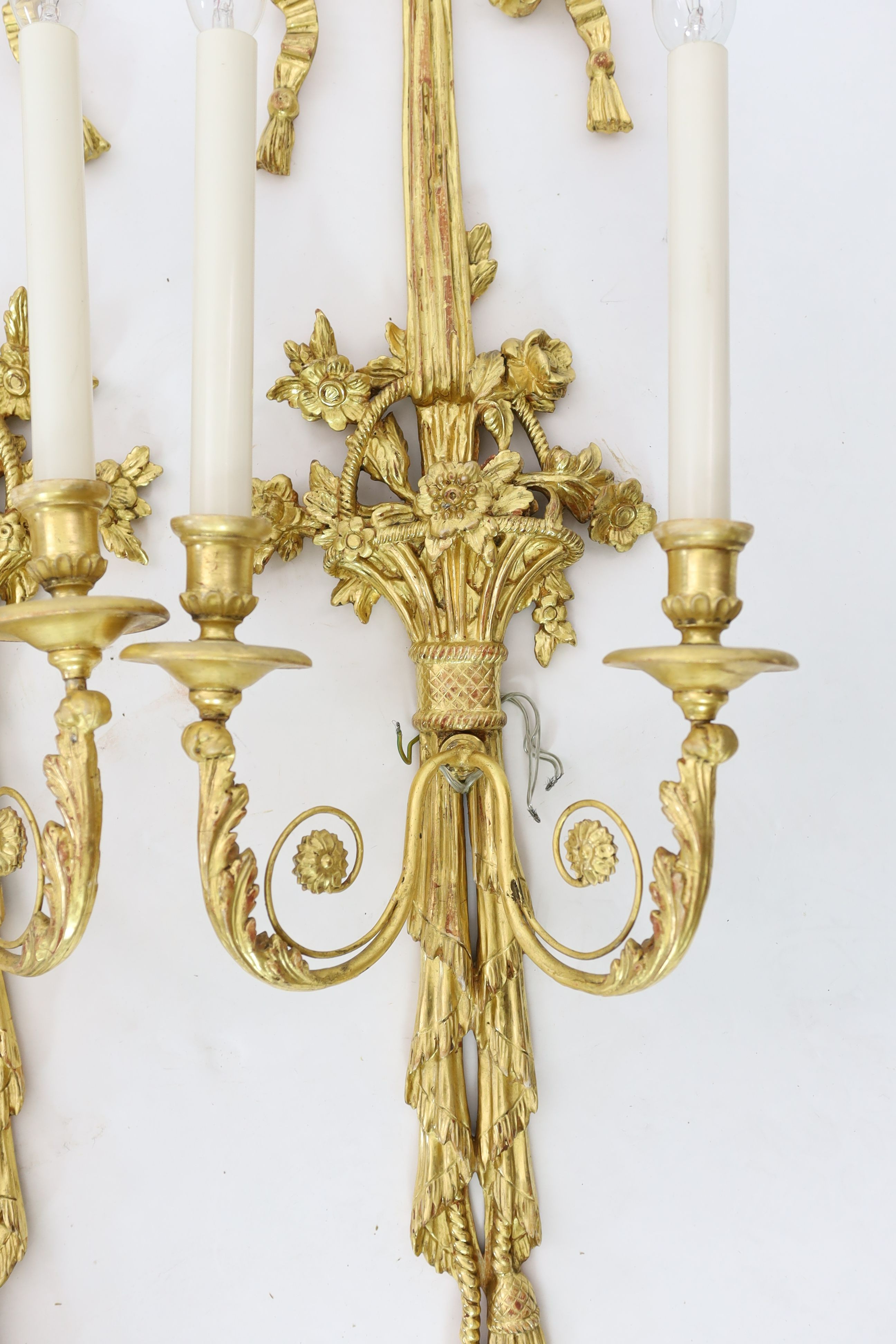 A pair of Louis XVI style carved giltwood wall lights, with ribbon crests and flowers in basket stems, over tasselled bases, height 85cm